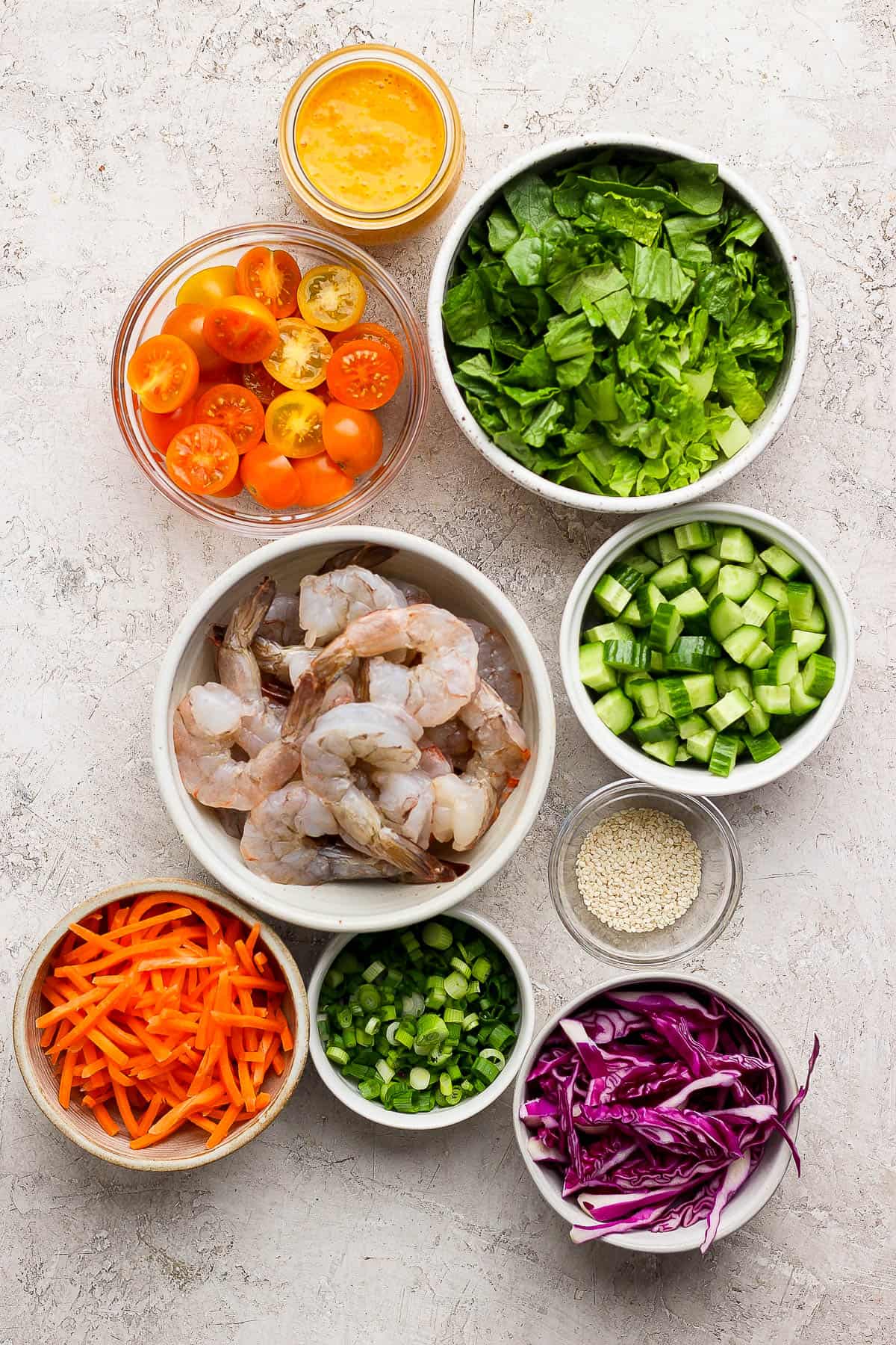 Individual bowls of carrot ginger dressing, halved cherry tomatoes, chopped romaine, raw shrimp, sliced and quartered mini cucumbers, sesame seeds, matchstick carrots, sliced green onion, and purple cabbage.