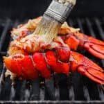 Lobster tails being grilled with someone brushing garlic herb butter over top.