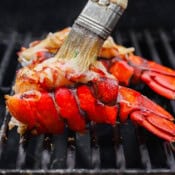 Lobster tails being grilled with someone brushing garlic herb butter over top.