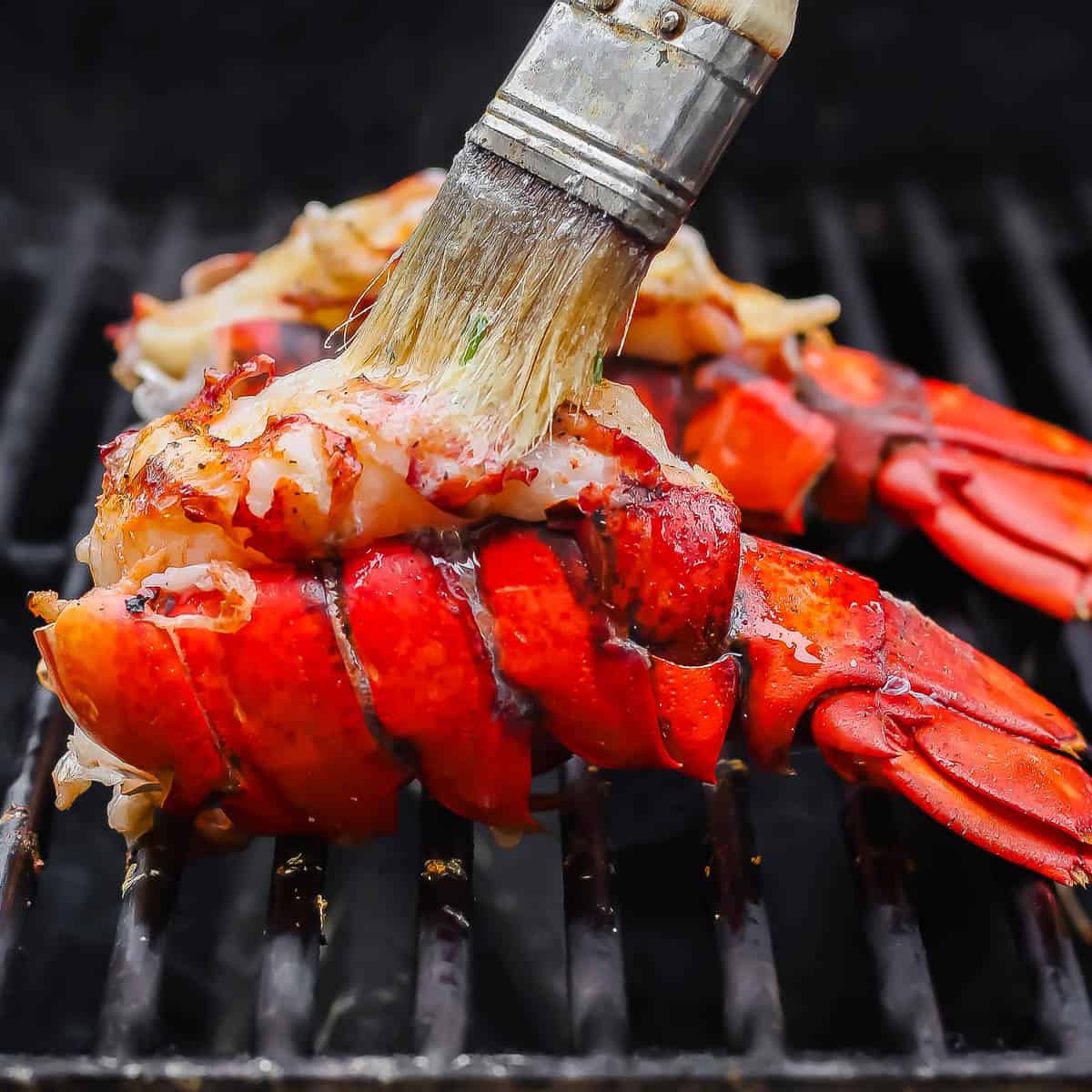 Lobster tails being grilled with someone brushing garlic herb butter over top.