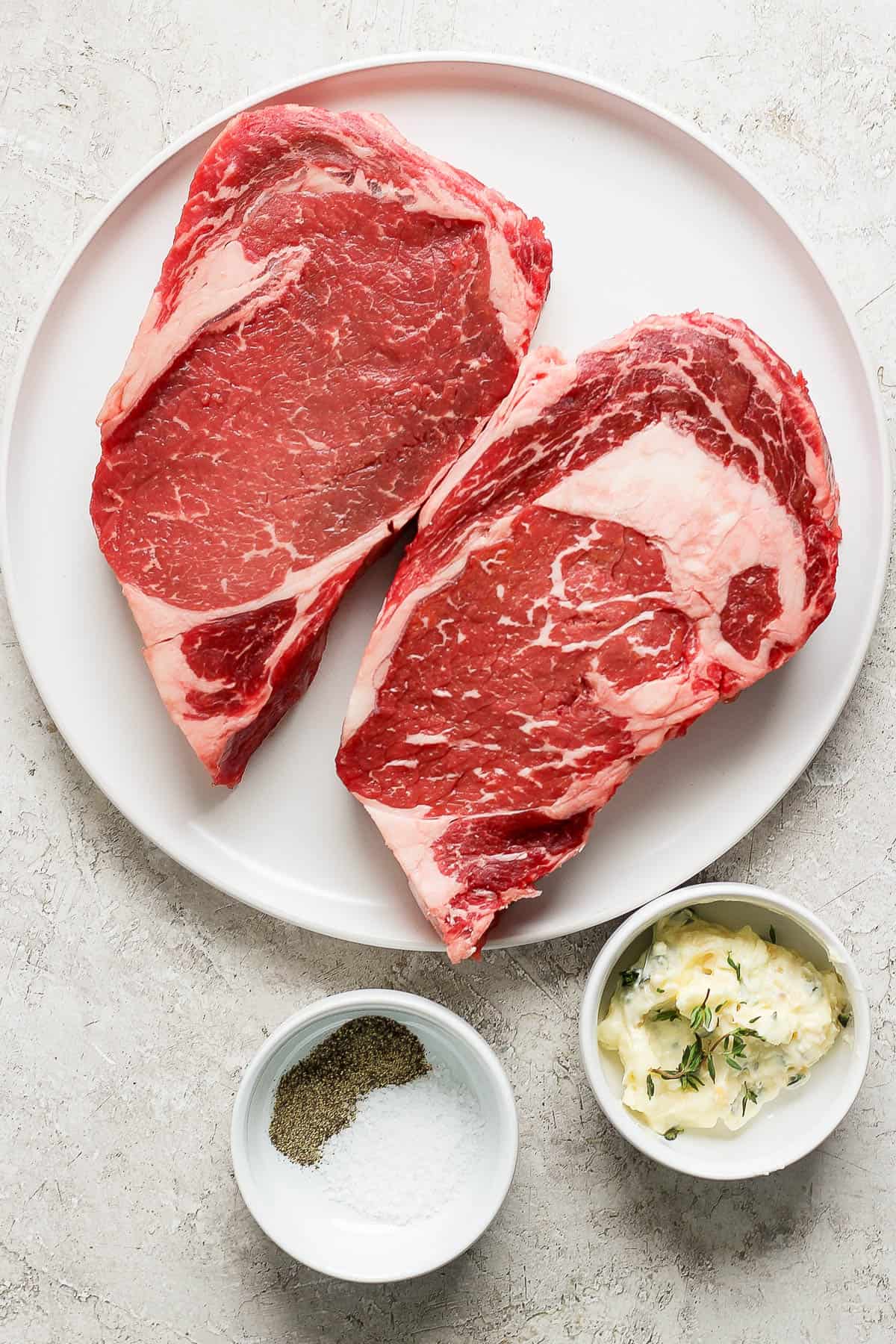 A plate with two ribeye steaks and a bowl of herbed butter and another bowl with salt and pepper.