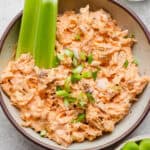 A bowl of high protein buffalo chicken dip with sliced green onions on top and celery stalks sticking out.