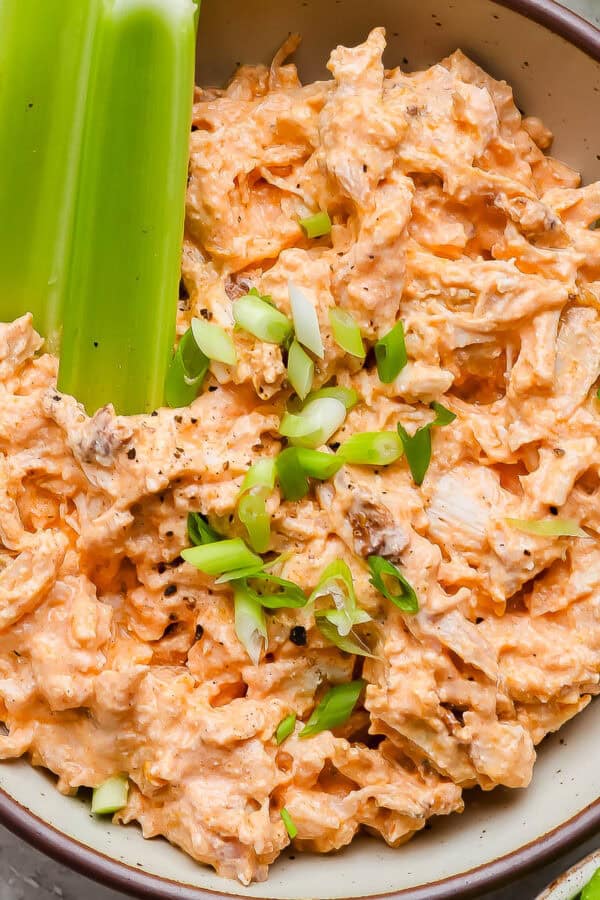 A bowl of high protein buffalo chicken dip with sliced green onions on top and celery stalks sticking out.