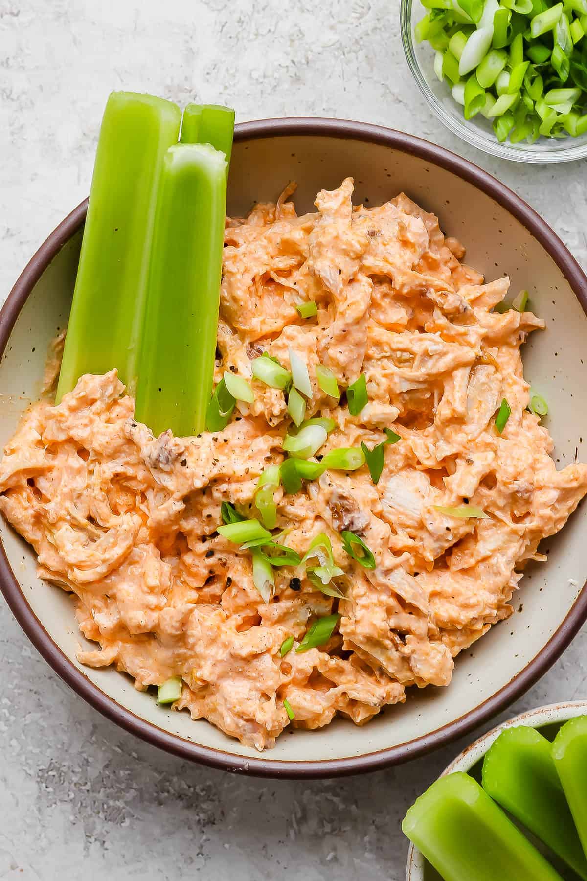 High protein buffalo chicken dip in a bowl with three celery stalks on the side. All topped with sliced green onions.