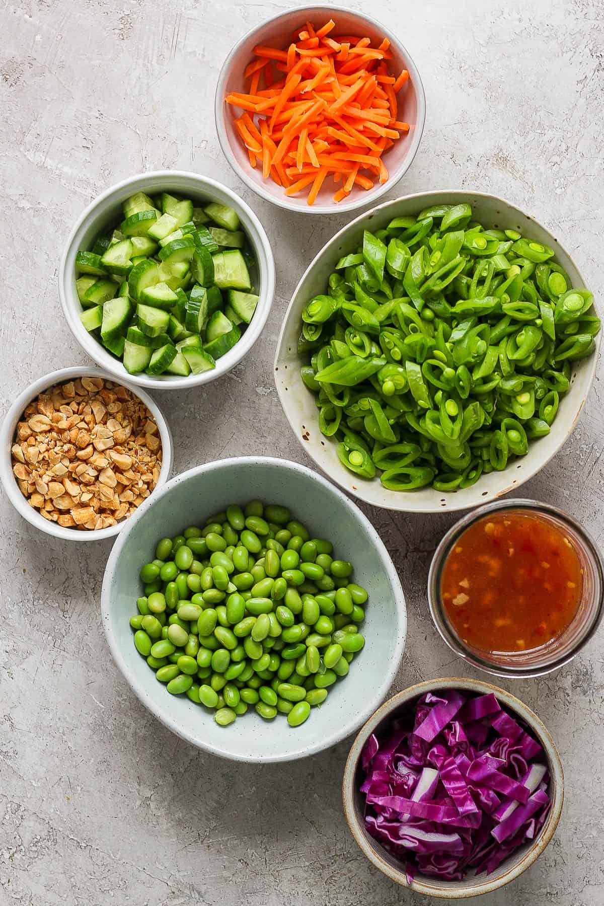 Individual bowls of matchstick carrots, sliced and quartered cucumbers, chopped peanuts, sliced snap peas, edamame, sliced purple cabbage, and sweet chili dressing.