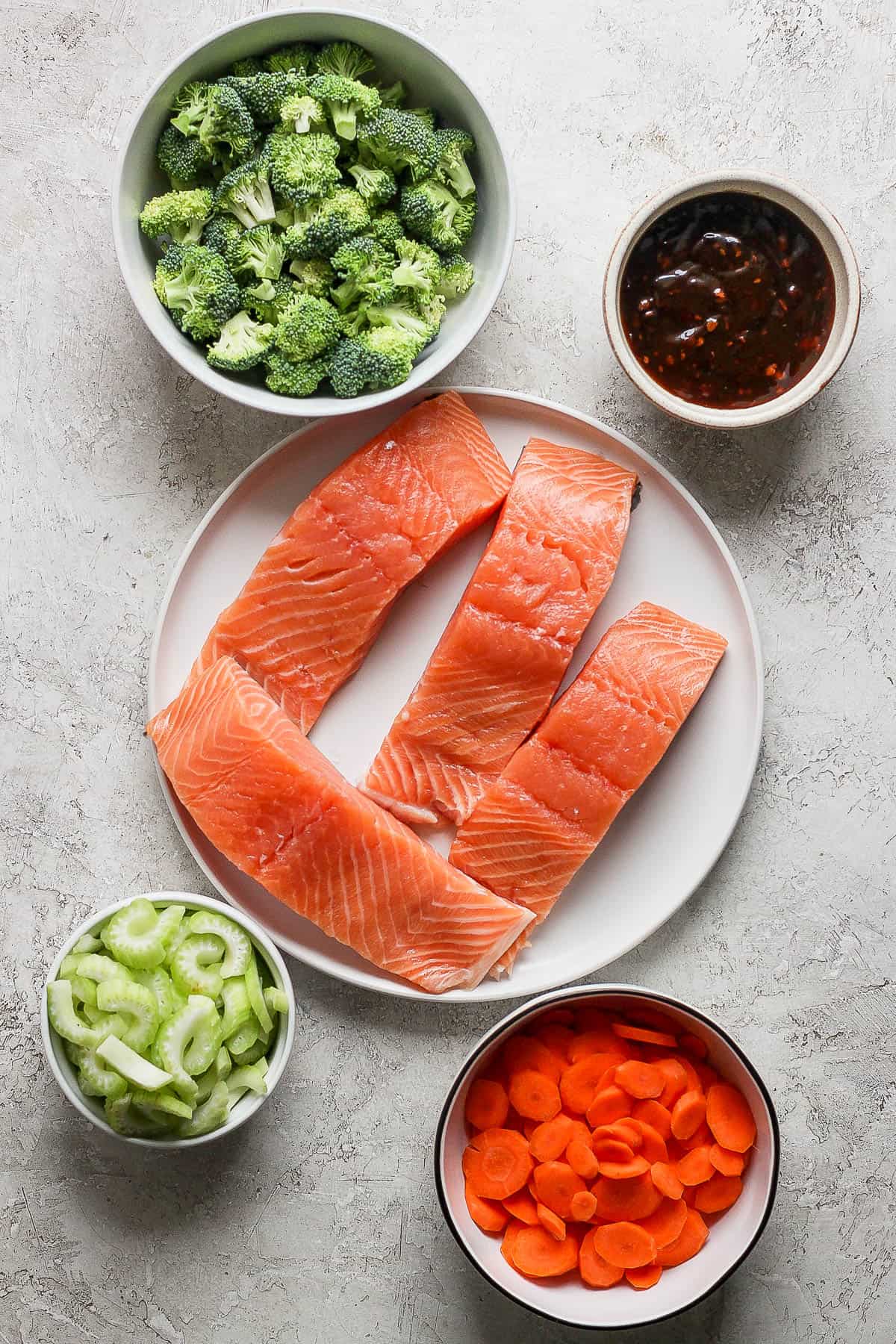 Individual bowls of chopped broccoli, sliced celery, and sliced carrots. A plate with 4 salmon filets and a small bowl of teriyaki sauce.