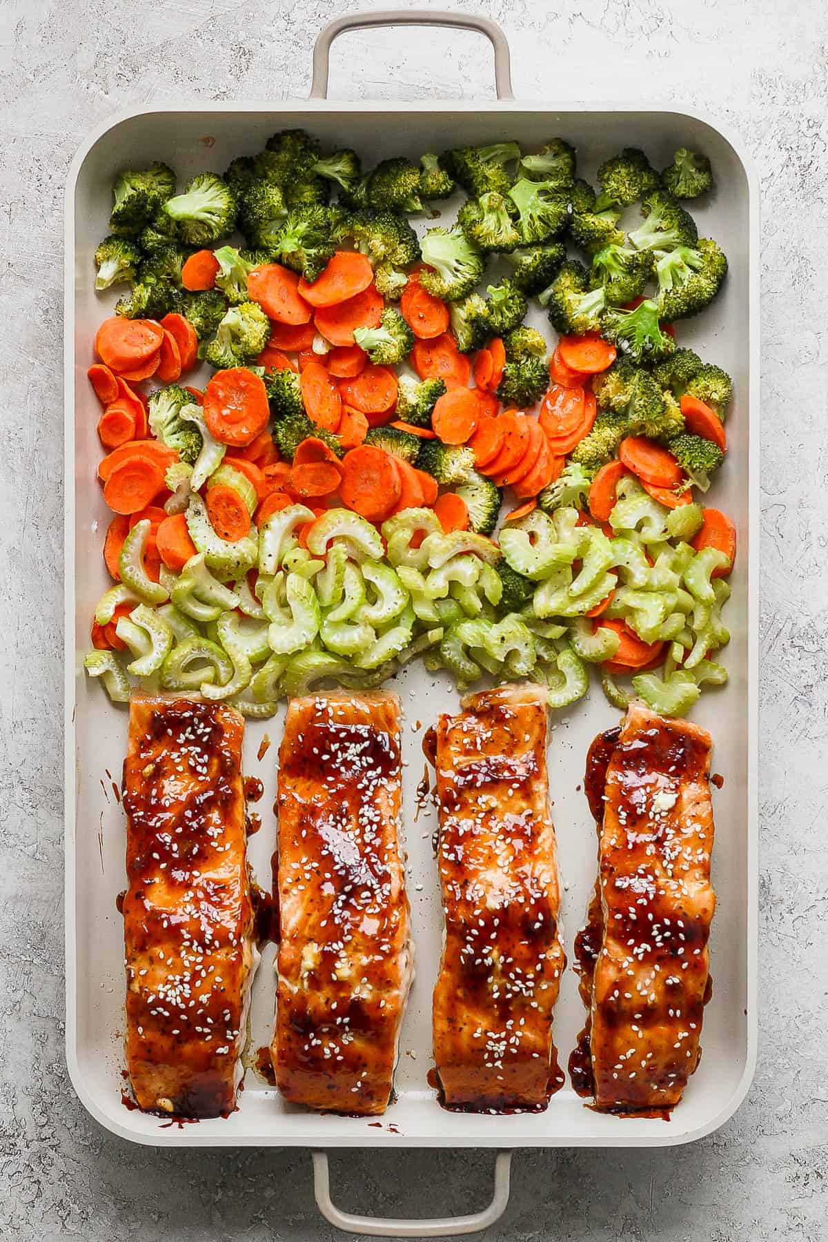 A shallow sheet pan with roasted broccoli florets, sliced carrots, sliced celery and 4 baked salmon filets topped with teriyaki sauce and sesame seeds.