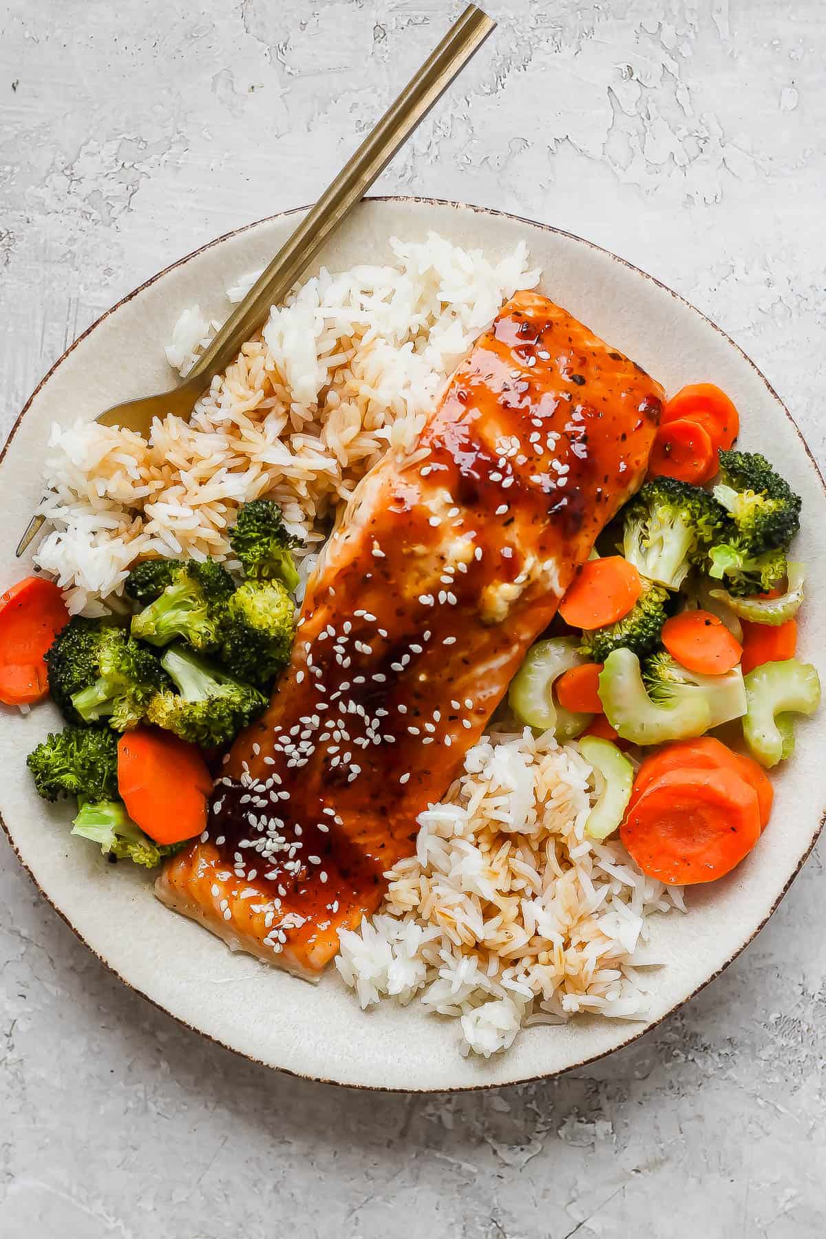 A filet of teriyaki salmon ton top of a bed of rice with cooked veggies on either side.