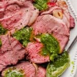 Sliced smoked leg of lamb on a plate with homemade chimichurri sauce over top and a spoon on the side.