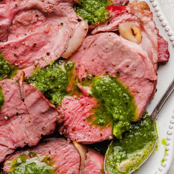 Sliced smoked leg of lamb on a plate with homemade chimichurri sauce over top and a spoon on the side.