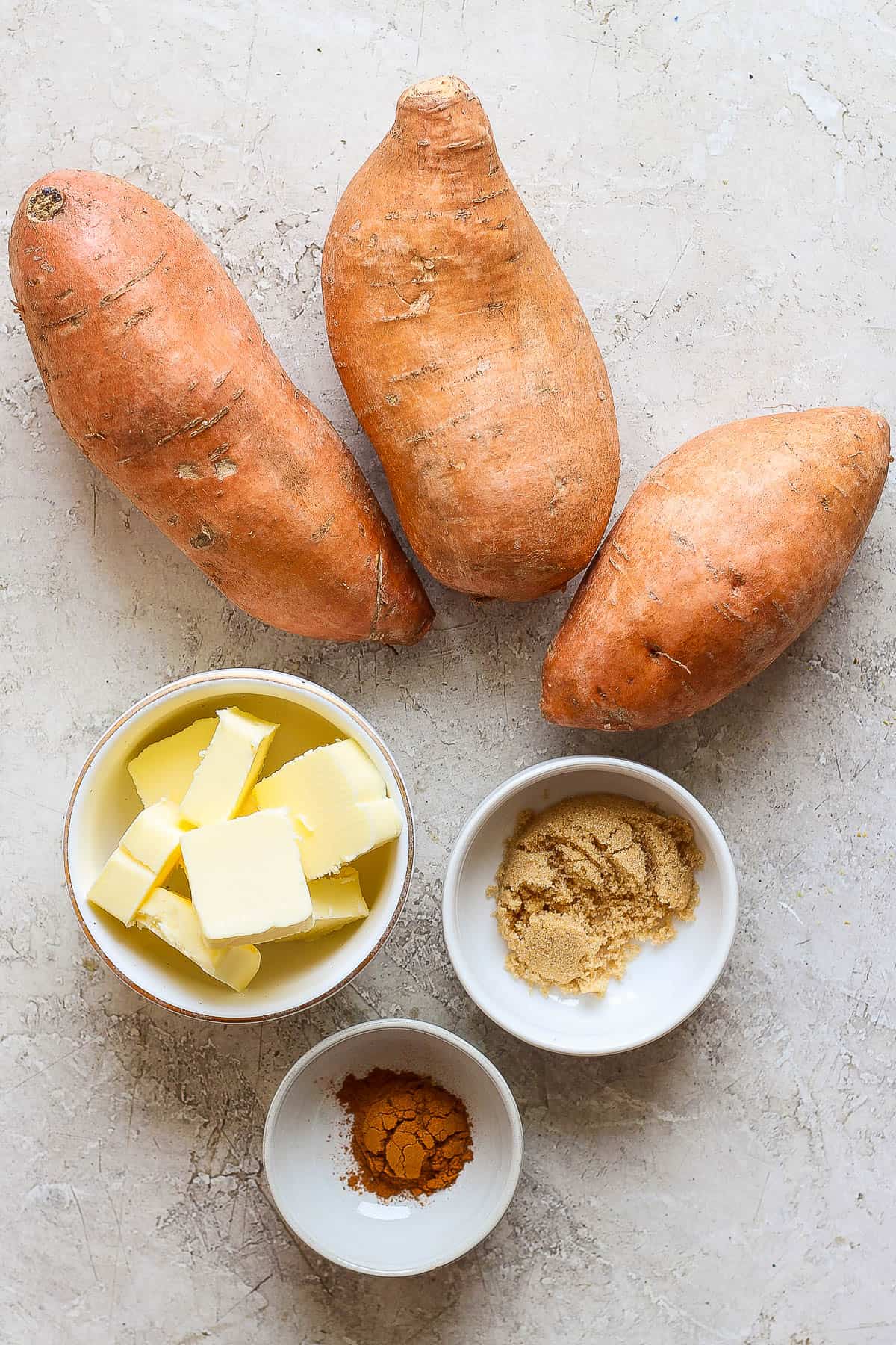 Three whole sweet potatoes and individual bowls of sliced butter, brown sugar, and all spice on the counter.