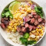 A bowl of rice topped with grilled steak and grilled street corn salad with lime wedges and fresh cilantro.