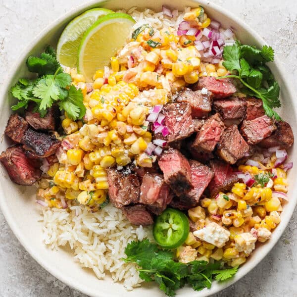 A bowl of rice topped with grilled steak and grilled street corn salad with lime wedges and fresh cilantro.