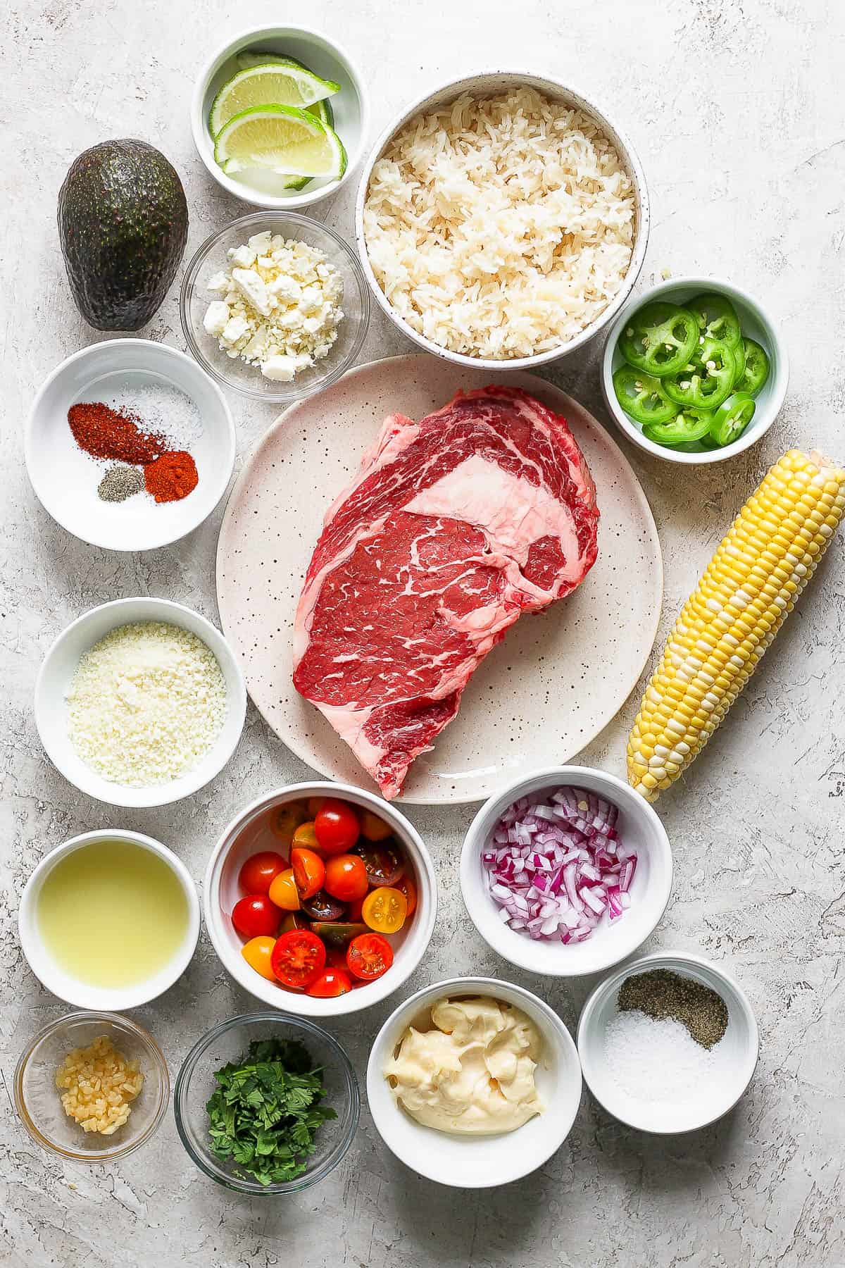 Individual bowls of lime wedges, cojita cheese, cooked white rice, jalapeno slices, seasonings, a full ribeye steak, feta cheese, corn on the cob, halved cherry tomatoes, olive oil, diced red onion, lime juice, minced garlic, chopped cilantro, mayo, and salt and pepper.