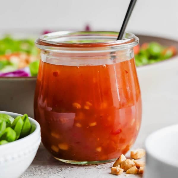 A jar of homemade sweet chili vinaigrette dressing with a spoon sticking out of it and a salad behind it.