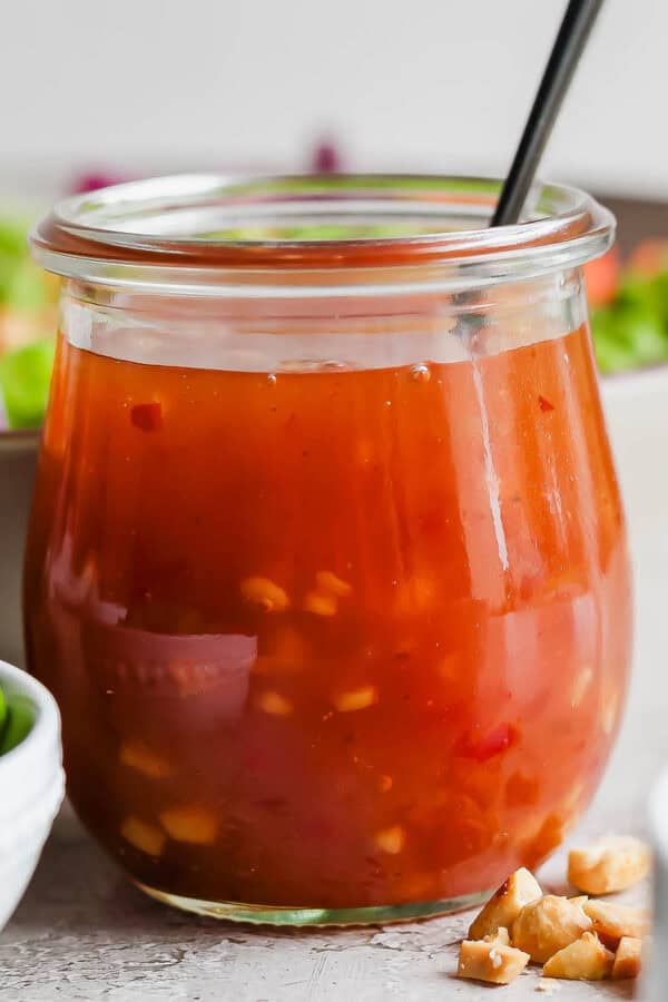 A jar of homemade sweet chili vinaigrette dressing with a spoon sticking out of it and a salad behind it.