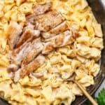 A cast iron skillet filled with creamy alfredo tortellini with chicken and a spoon sticking out.