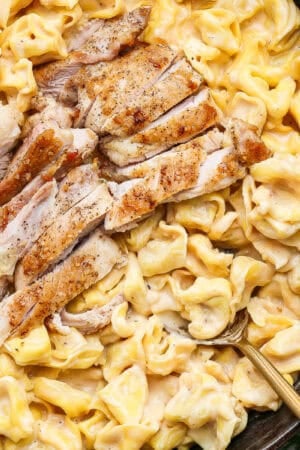 A cast iron skillet filled with creamy alfredo tortellini with chicken and a spoon sticking out.