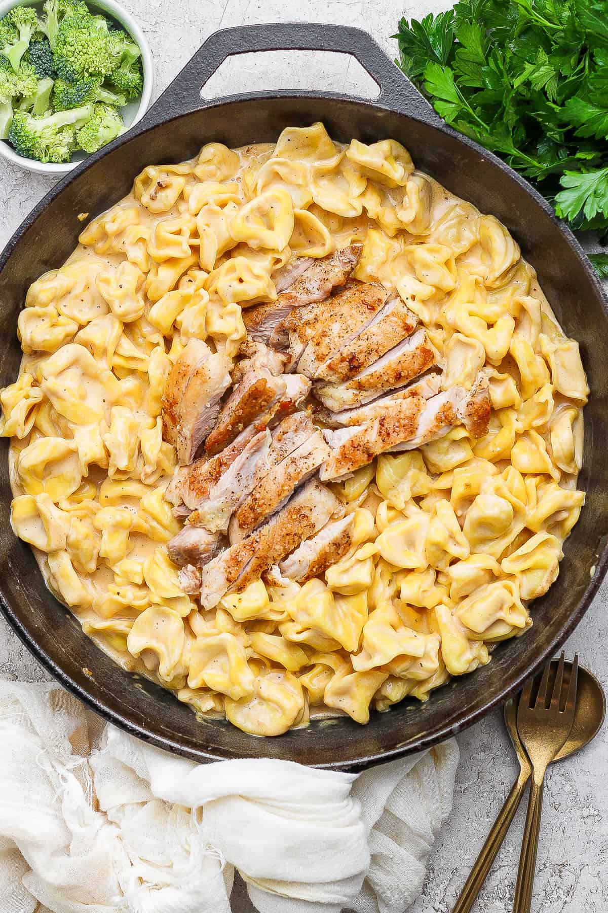 Alfredo tortellini in a cast iron skillet with sliced chicken on top and next to a small bowl of broccoli and bunch of fresh parsley.