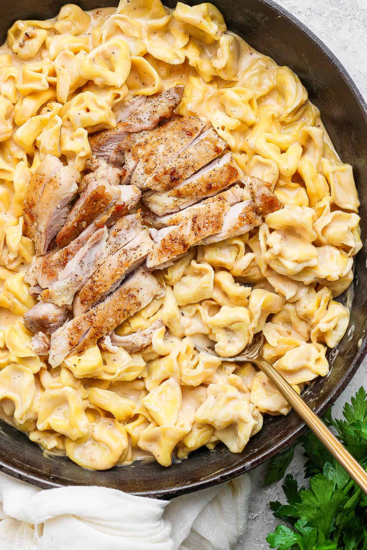 Sliced chicken thighs on top of tortellini alfredo in a cast iron skillet.