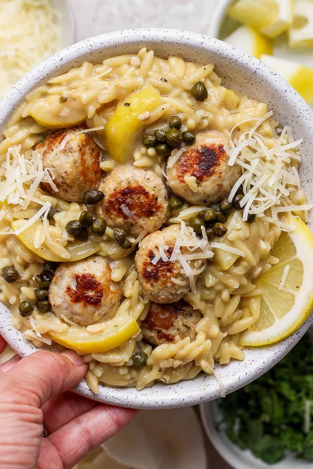 A hand holding a bowl of chicken piccata meatballs with orzo and parmesan on top.