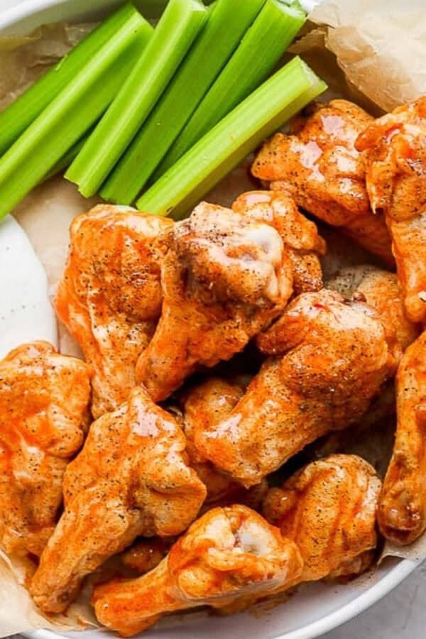 Several buffalo chicken wings piled in a bowl with ranch dressing and celery sticks that's lined with parchment paper.