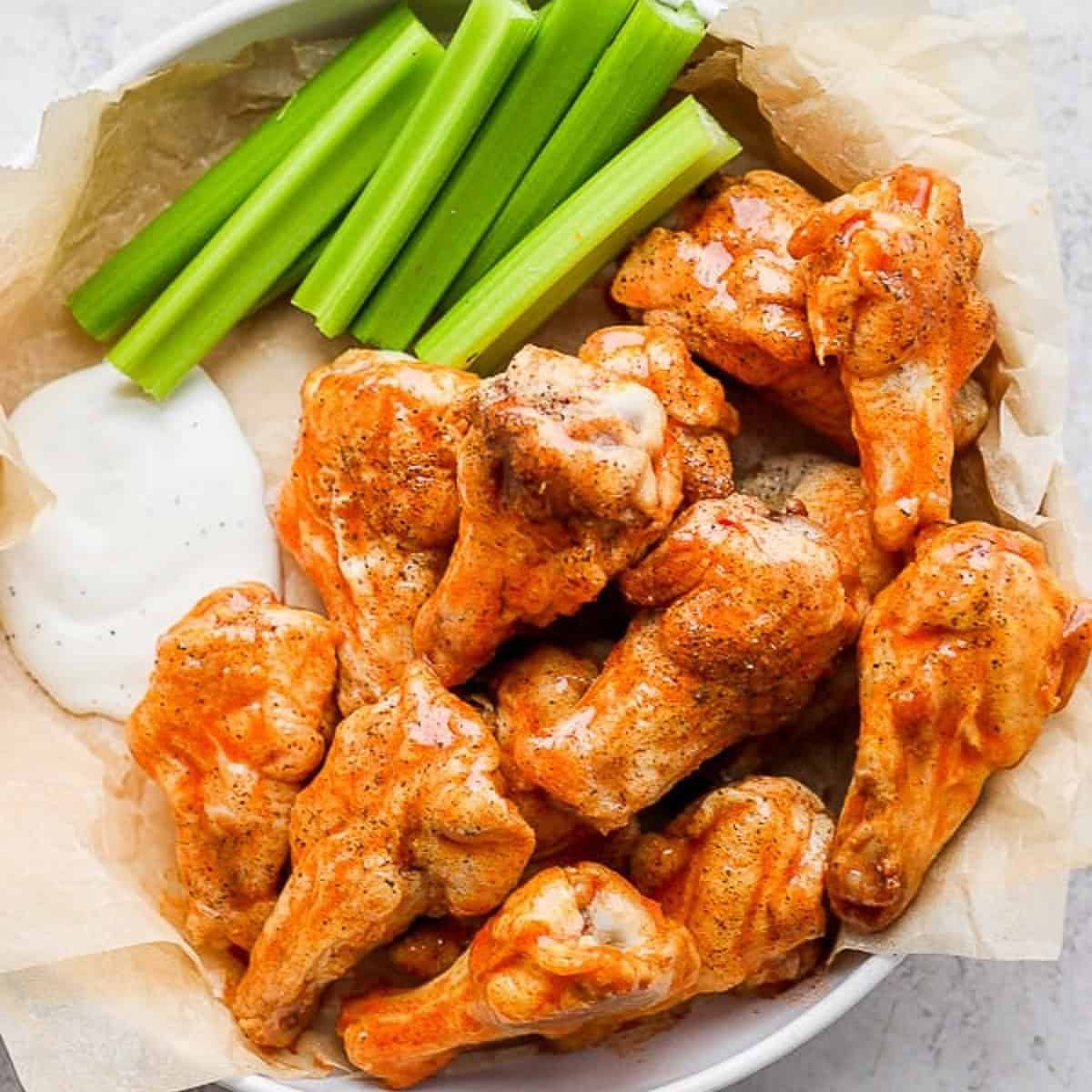 Several buffalo chicken wings piled in a bowl with ranch dressing and celery sticks that's lined with parchment paper.