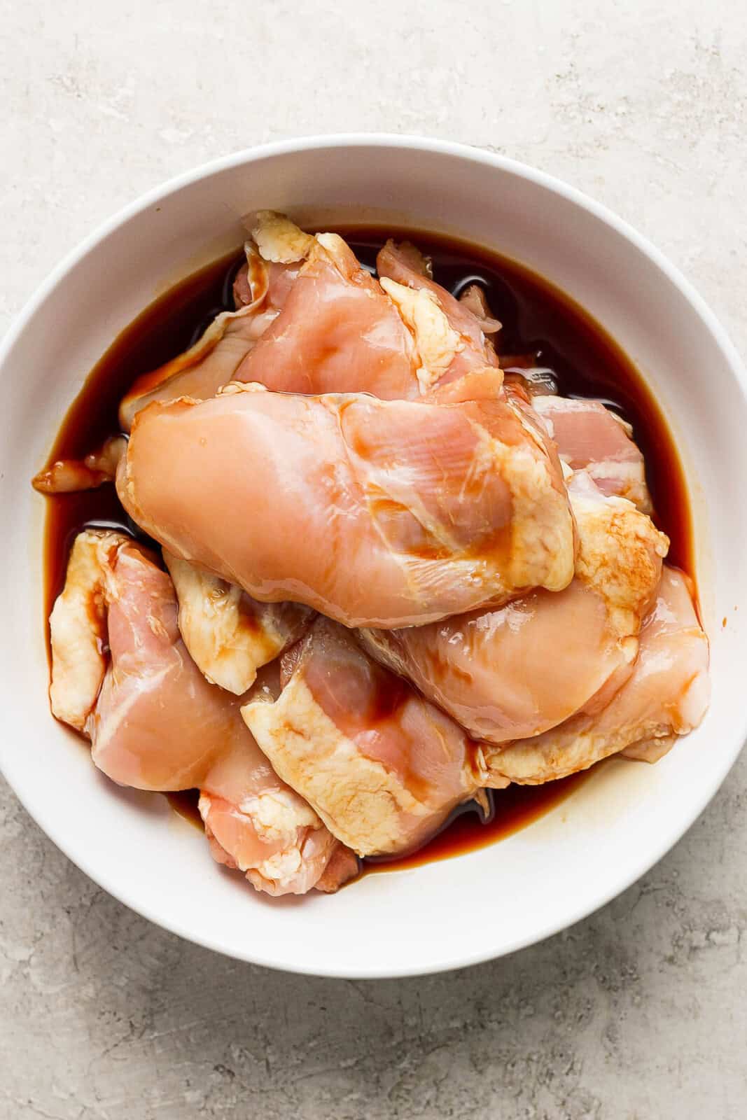 Chicken thighs marinating in soy sauce in a white bowl.