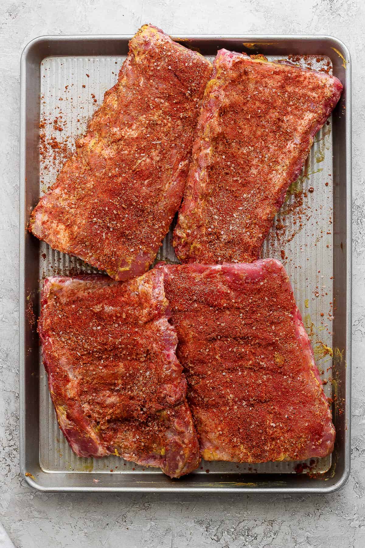 Half racks of ribs covered in dry rub on a sheet pan.