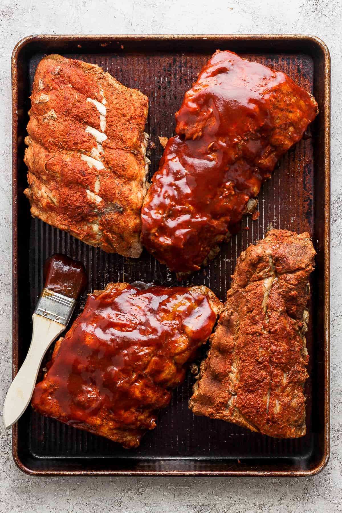 Half of the cooked ribs brushed with bbq sauce and half do not have sauce on a sheet pan.