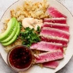 Slices of seared ahi tuna on a plate with crispy rice, sliced avocado, spicy mayo, cilantro, and a dish of sauce for dipping.