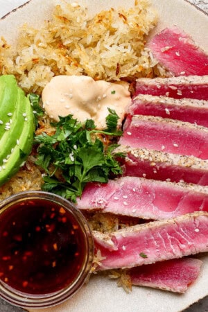 Slices of seared ahi tuna on a plate with crispy rice, sliced avocado, spicy mayo, cilantro, and a dish of sauce for dipping.
