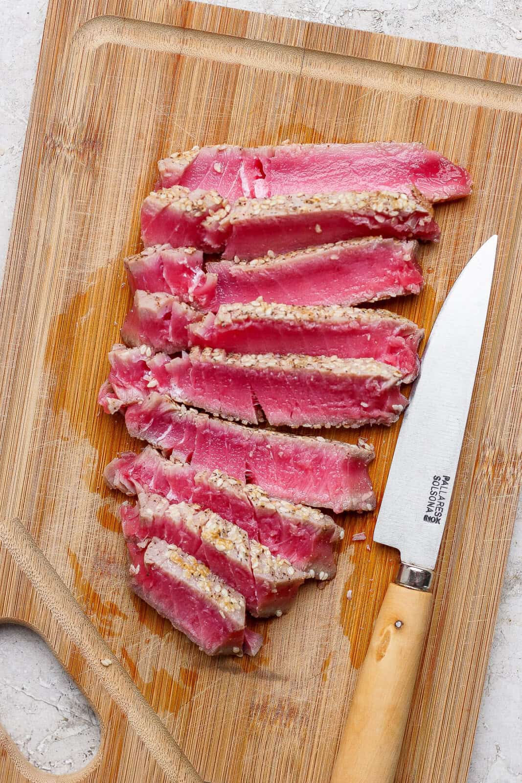 A seared tuna steak cut into slices on a cutting board with a knife on the side.