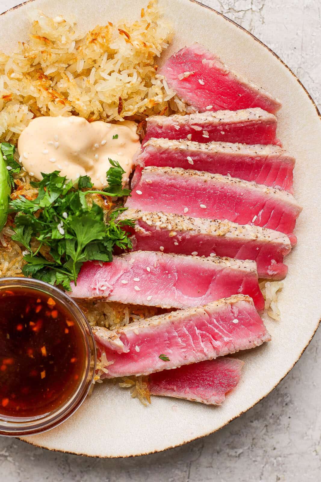 Slices of seared ahi tuna on a plate with crispy rice, fresh cilantro, spicy mayo, and a dish of dipping sauce.