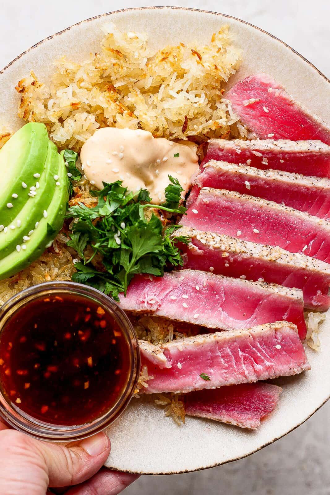 Slices of seared ahi tuna on a plate with crispy rice, slices of avocado, spicy mayo, and a small dish of dipping sauce.