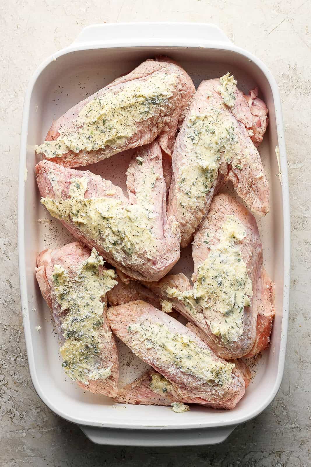Herbed butter spread on raw turkey wings in a white baking dish.