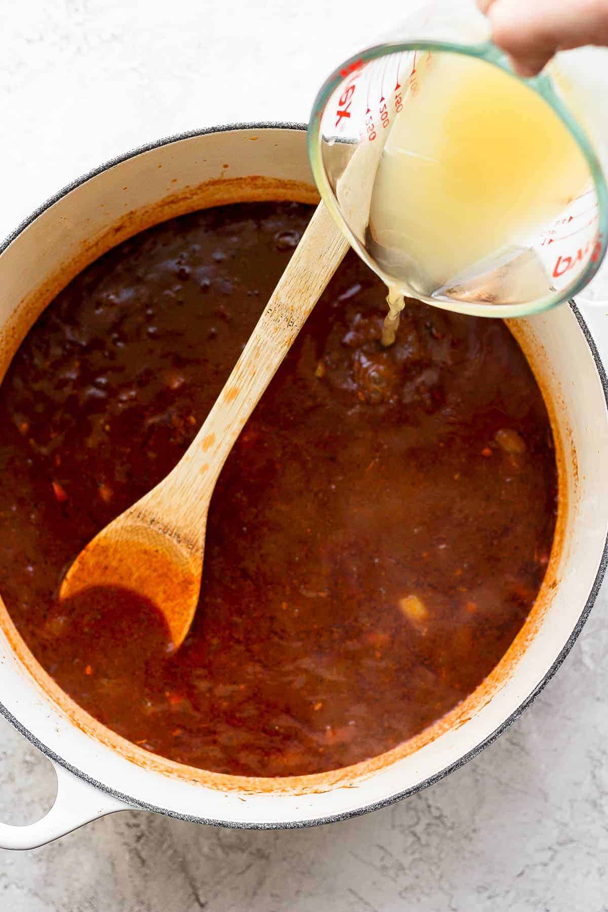 Chicken broth being poured in to a pot of chicken enchilada soup with a wooden spoon in it.