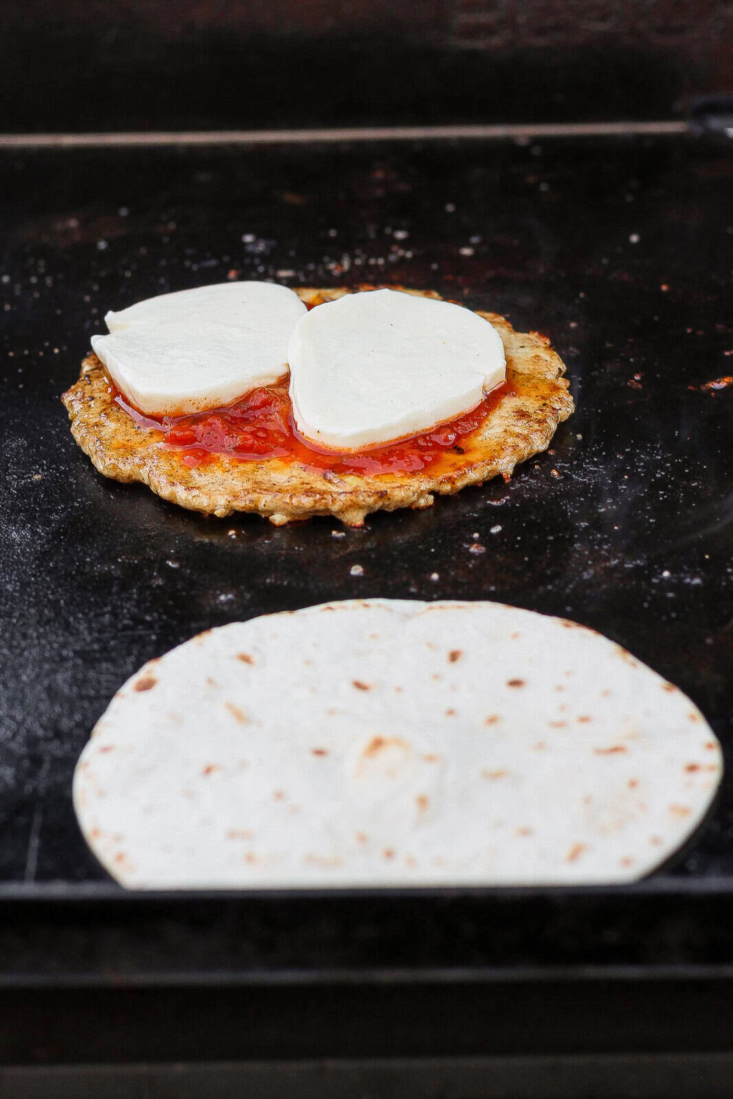 A hot Blackstone griddle with a smashed chicken burger covered in marinara sauce and two slices of cheese next to a tortilla.
