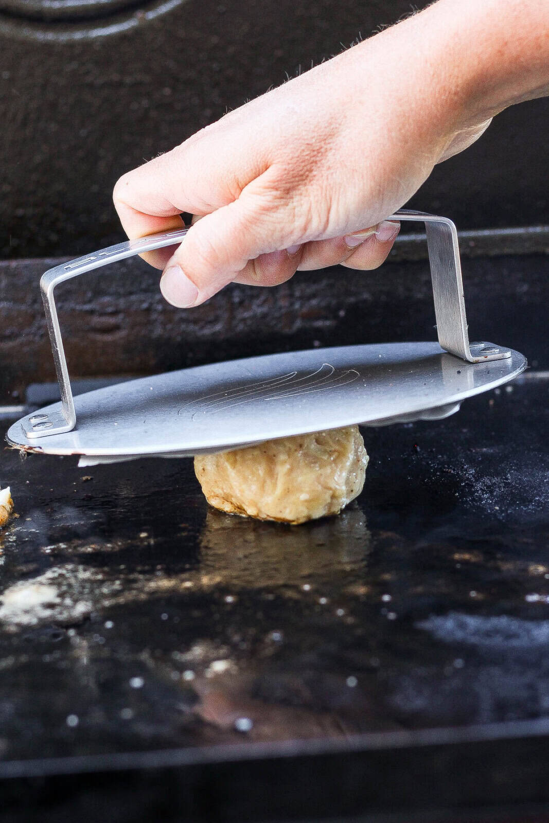 A hand pressing a chicken burger patty flat with a burger press and piece of parchment paper on a hot griddle.