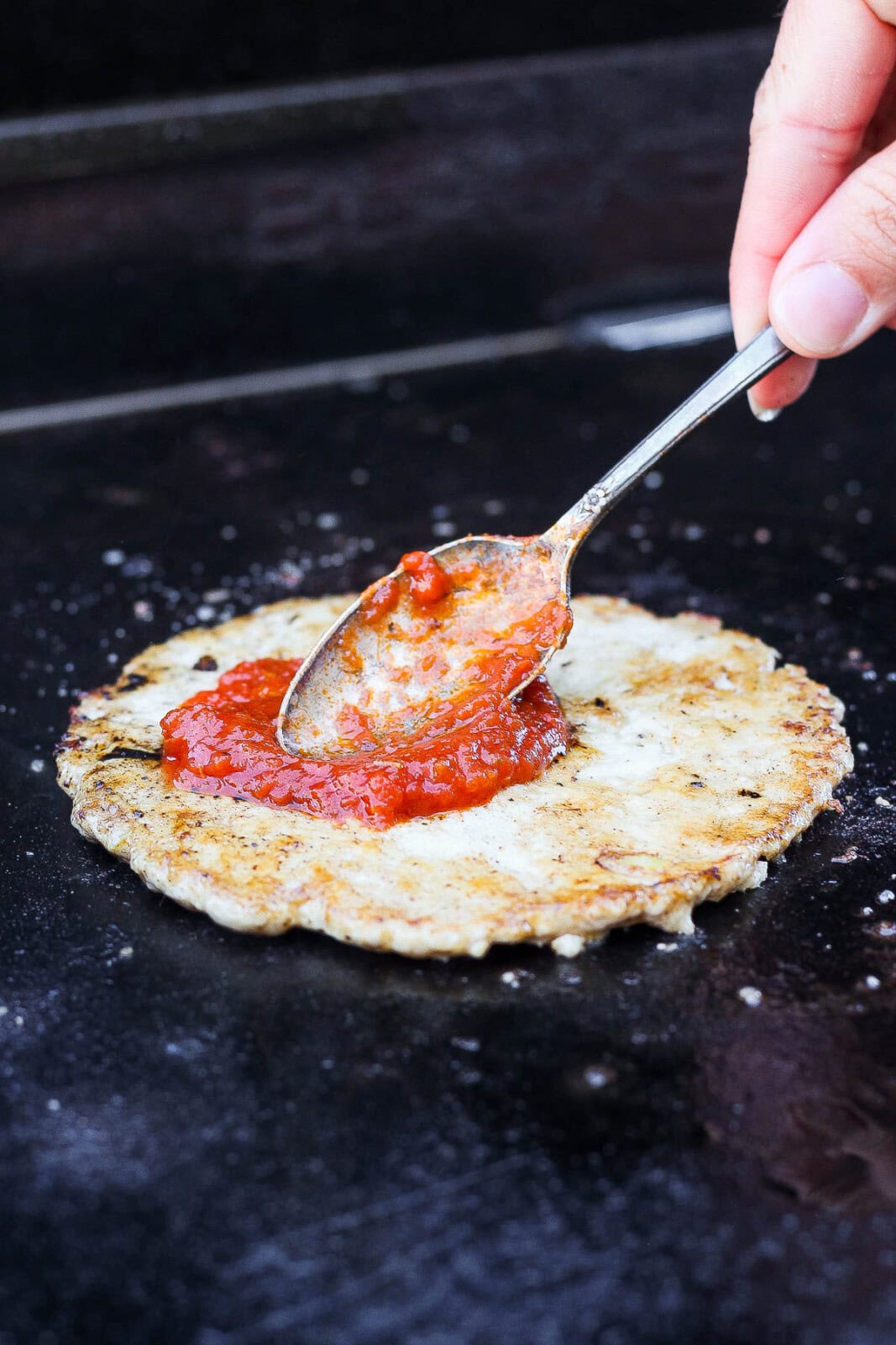 A hand using a spoon to spread marinara sauce on a smashed burger patty on the Blackstone.