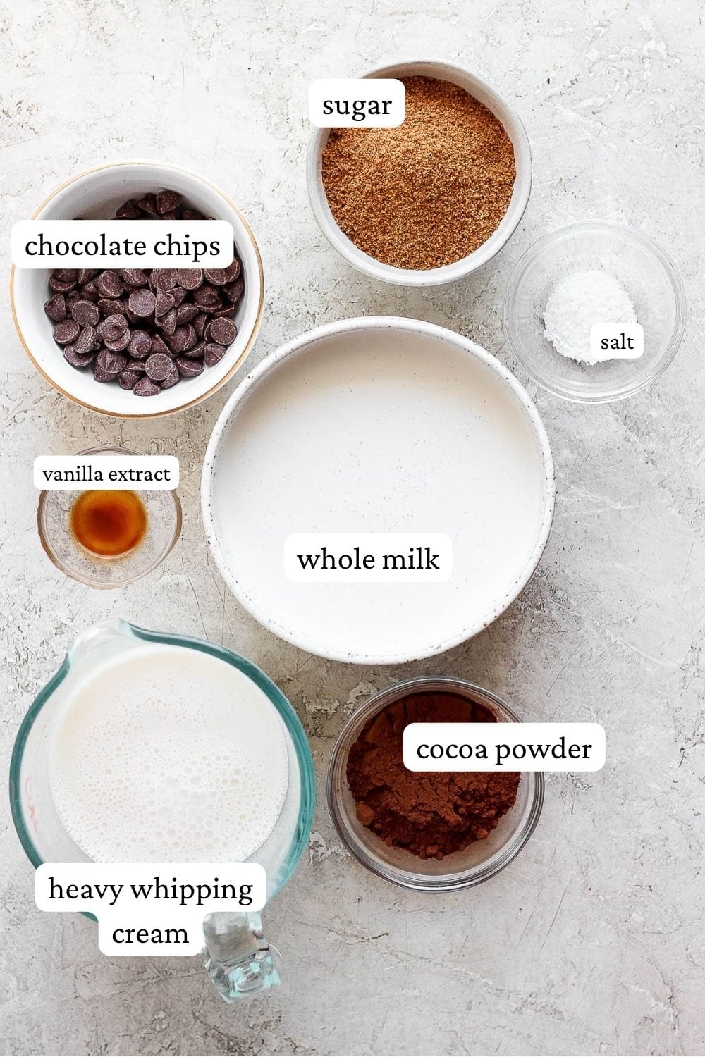 Individual bowls of sugar, chocolate chips, salt, whole milk, vanilla extract, heavy whipping cream, and cocoa powder on a counter top.