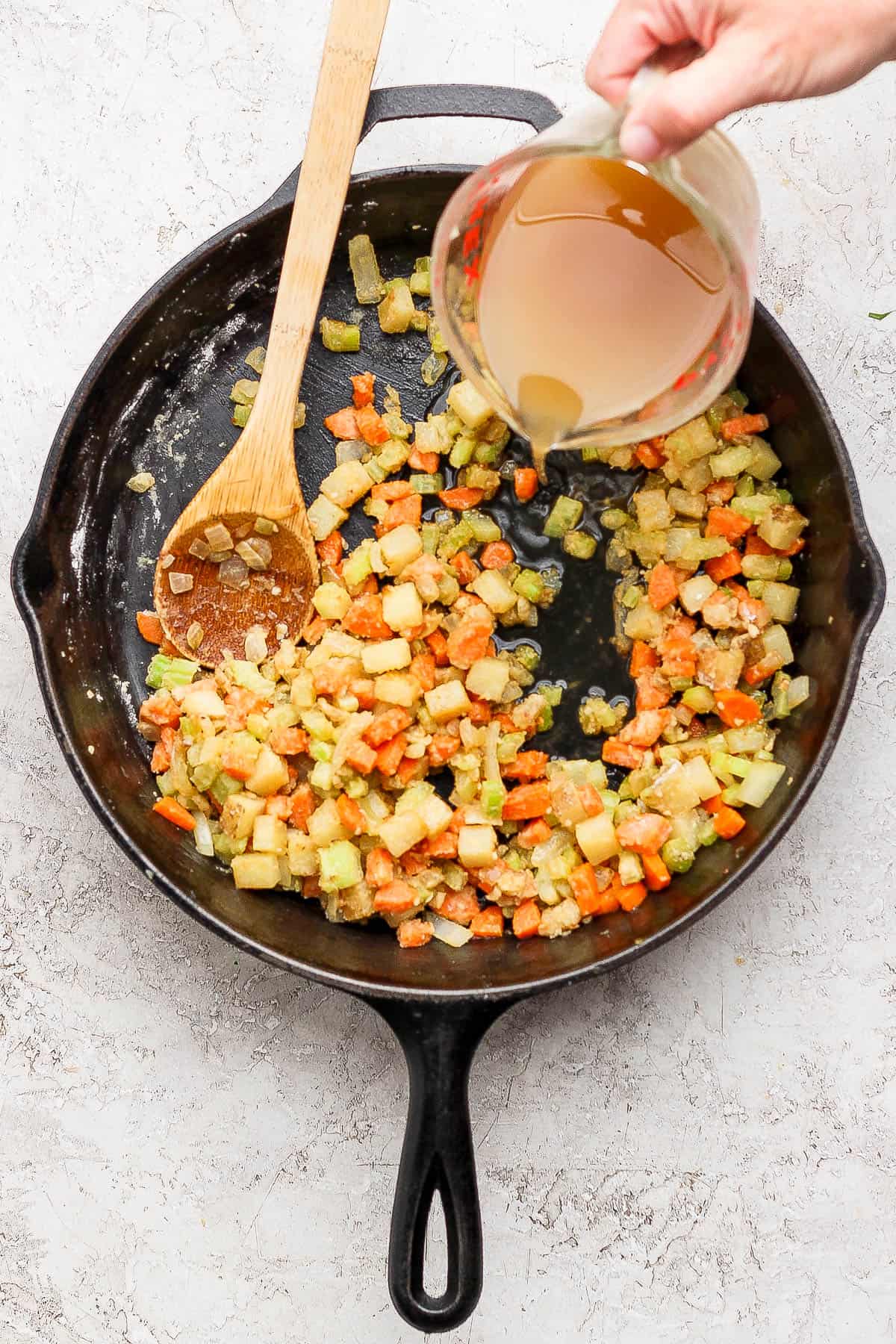 Sautéd garlic, onion, carrots, and celery in a skillet coated with flour and a hand pouring chicken broth over the top.