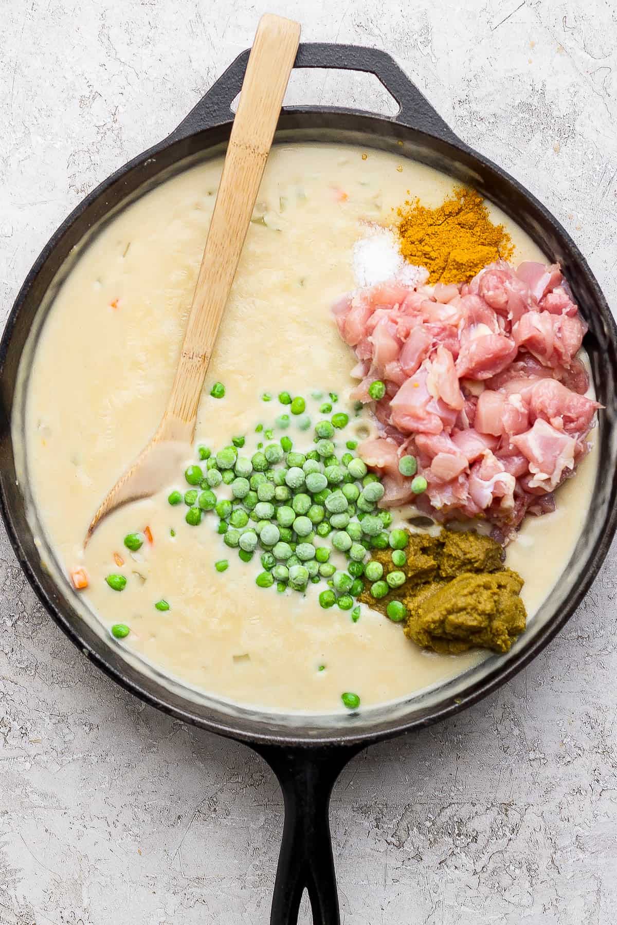 Creamy pot pie filling in a skillet with frozen peas, raw chicken, and seasonings.