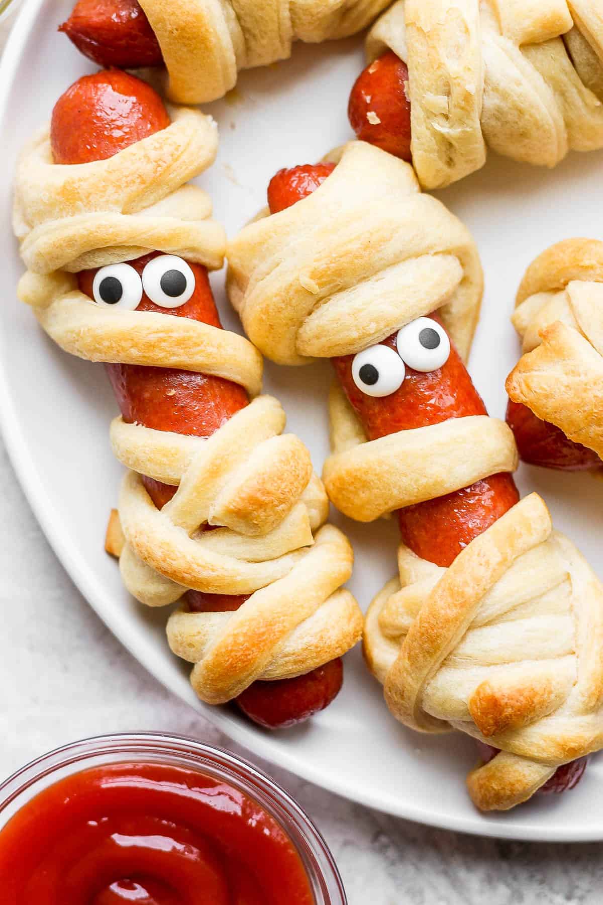 Several mummy hot dogs with candy eyes on a white plate.