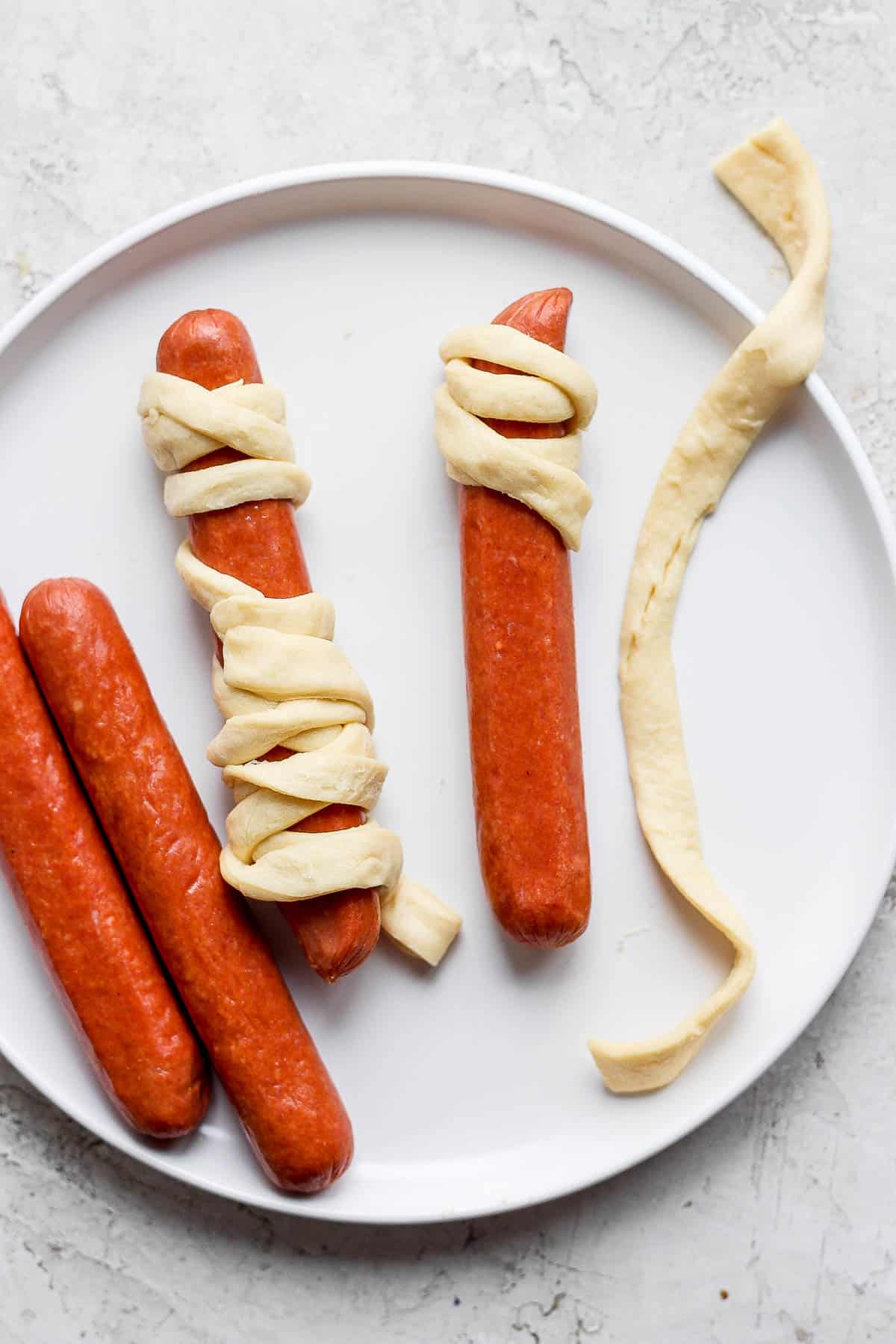 Four hot dogs on a white plate, two have some crescent dough strips wrapped around them and two do not.