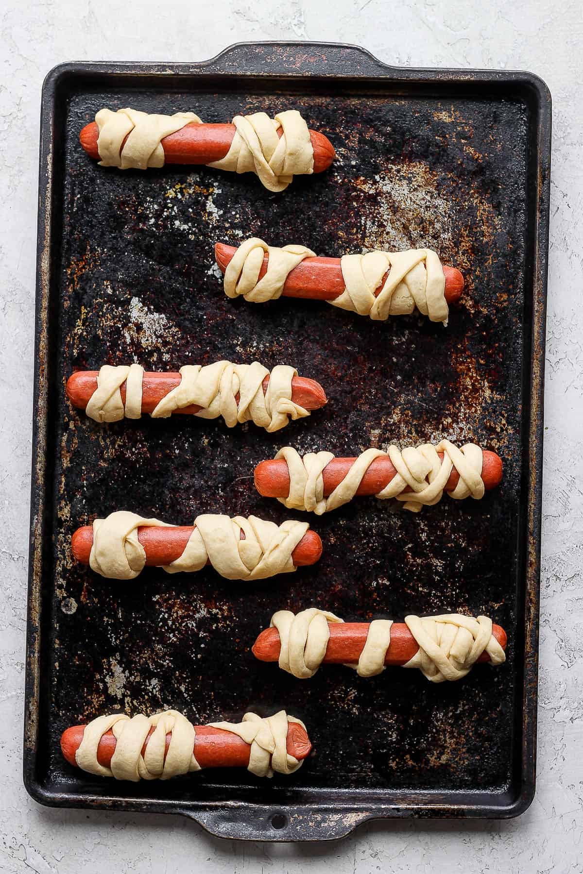 Seven hot dogs wrapped in crescent roll dough strips on a baking sheet before baking.
