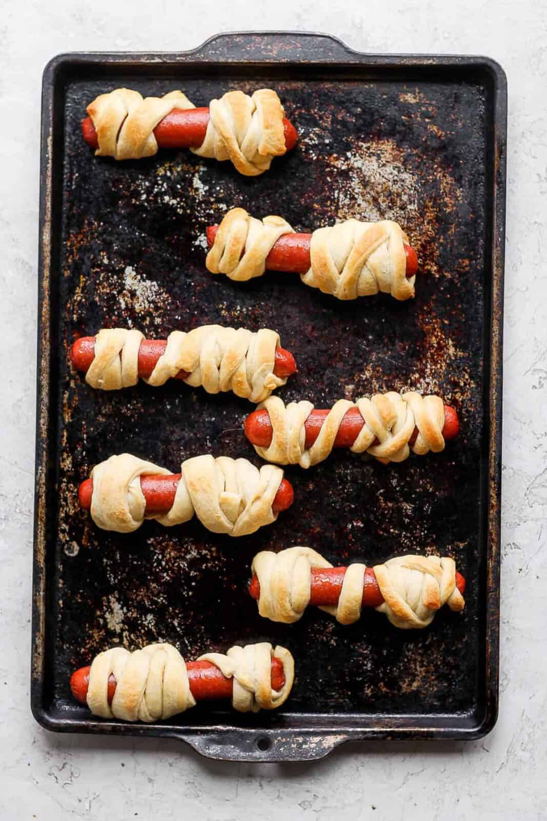 Seven hot dogs wrapped in crescent roll dough strips on a baking sheet after baking.