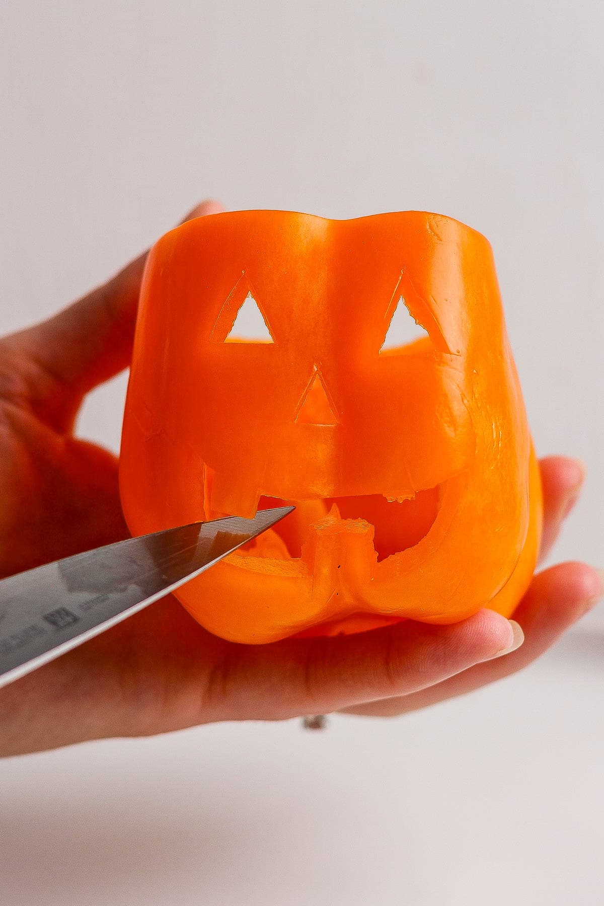 One hand holding an orange bell pepper and the other hand is using a paring knife to cut a jack-o-lantern face in the side of the pepper.