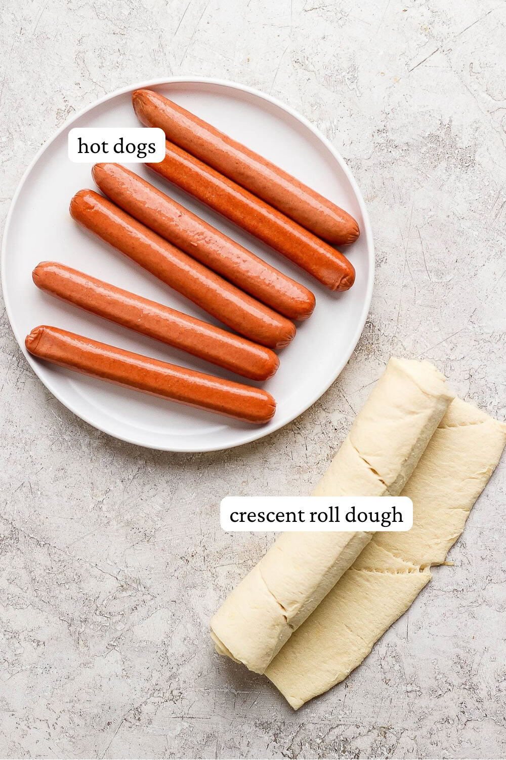 Raw hot dogs on a white plate next to a roll of crescent roll dough.