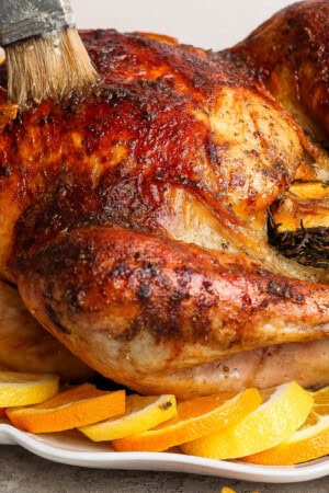 A fully cooked orange turkey on a platter with orange slices and being basted with a basting brush.